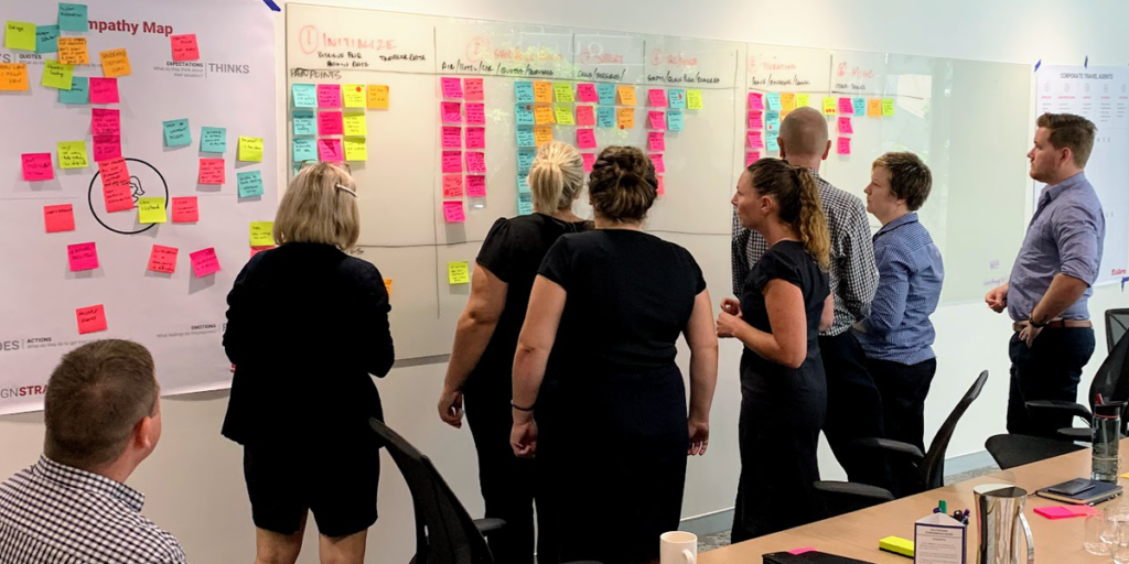 Photo of clients standing while examining the pain-points attached to the journey map hanging on the wall in a Design Thinking workshop